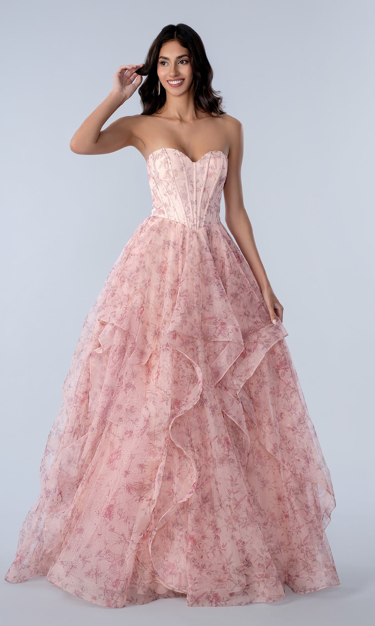 A woman models the Stella Couture 25119 Floral Prom Ball Gown, featuring a strapless corset bodice and a pink, floral-patterned glitter tulle skirt, against a plain light background.